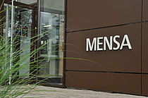 Der Eingang zur Mensa: Eine geöffnete Glastür, an der Wand rechts daneben steht in großen weißen Buchstaben"MENSA". Links ragen grüne Blätter ins Bild.