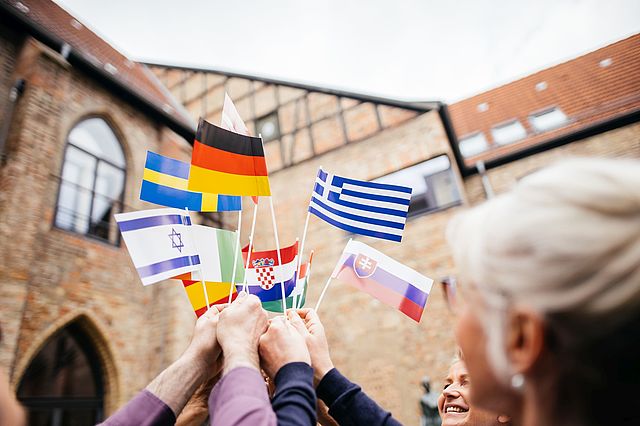 Mehrere Menschen halten kleine Flaggen verschiedener europäischer Staaten nach oben und zusammen. Im Hintergrund ein Fachwerkhaus mit gotischen Bögen.