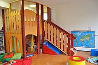 Day care center - playroom. In front left a cushion rope. A large wooden play structure with safety net. On the right a staircase leads up. At the back right a child-friendly world map on the wall.