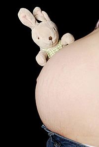 An unclothed baby belly from the left side against black background. On it sits a light brown plush bunny in white-green checkered clothes. 