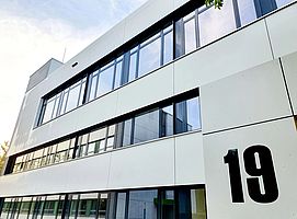 Das neue Lehrgebäude, Haus 19 (Ausschnitt). Ein modernes, weißes Gebäude mit großen Fenstern. Rechts eine große schwarze 19, die Gebäudenummer.