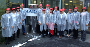 Vierzehn Personen, mit weißen, leicht transparenten Schutzkitteln und roten Helmen, im Hintergrund das Firmenlogo MARS