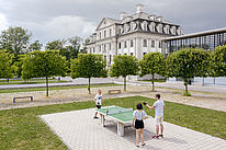 Students are playing table tennis. Behind them are trees, behind them a large white building.