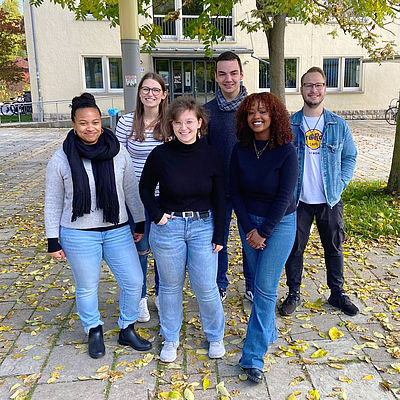 The six members of the student council for social management are standing on the square in front of the library. Behind them are two trees and a lantern, behind them is the entrance to the library. There are colourful leaves on the square.