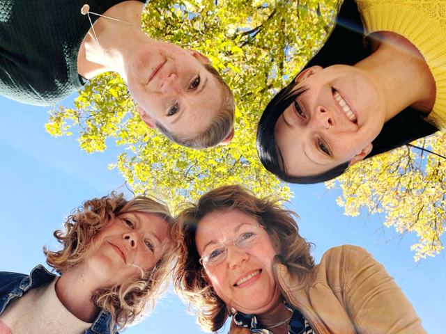 Die vier Mitglieder des Teams schauen fröhlich von oben nach unten in die Kamera. Im Hintergrund blauer Himmel und das gelbe Herbstlaub eines Baumes.