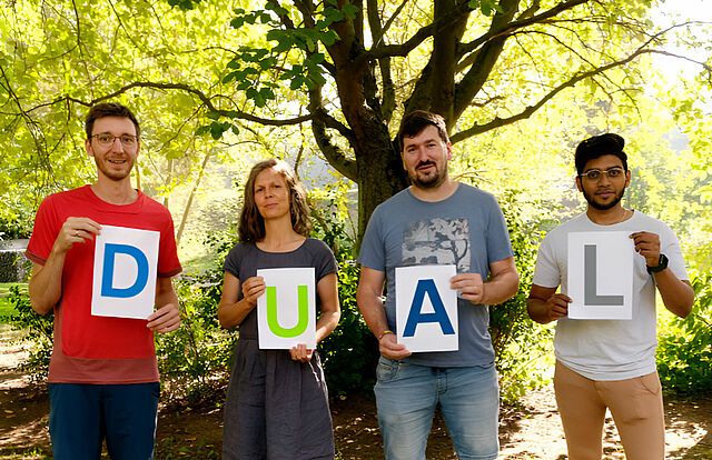 Vier Personen stehen nebeneinander. Jeder hält ein Schild mit einem Buchstaben hoch, zusammen ergibt es das Wort DUAL. Im Hintergrund stehen Bäume mit hellgrünem Laub.