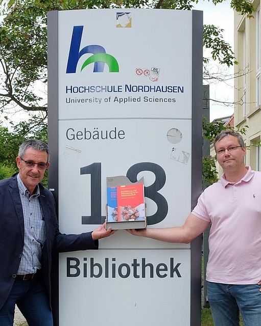 Professor Schwien und Daniel Hoffmeister mit ihrem neuen Buch vor dem Schild der Hochschulbibliothek