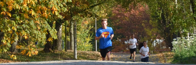 Campus run 2018, two runners run the course, on the left the brick building of AKI, in the background spectators, on the right the parking lot with some cars, behind it birch trees.