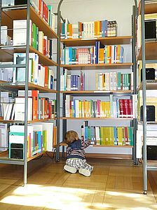 3 high bookshelves, arranged in a U. A toddler in plaid pants and a blue sweater crawls across the parquet floor. He reaches for a book with his right hand.