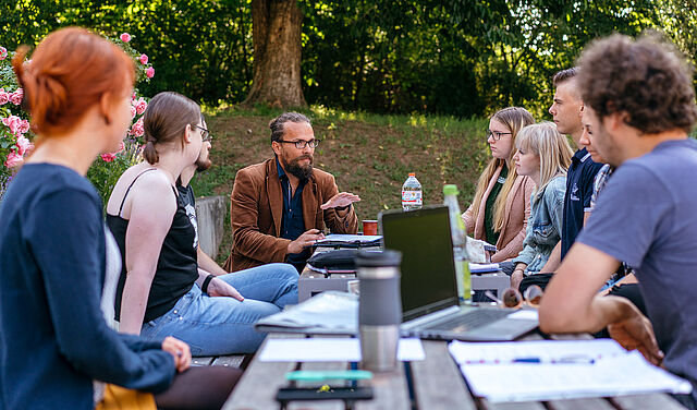 Studierende sitzen gemeinsam mit ihrem Professor an einem Tisch auf unserem grünen Campus. Der Prof. trägt Brille und Bart und gestikuliert.