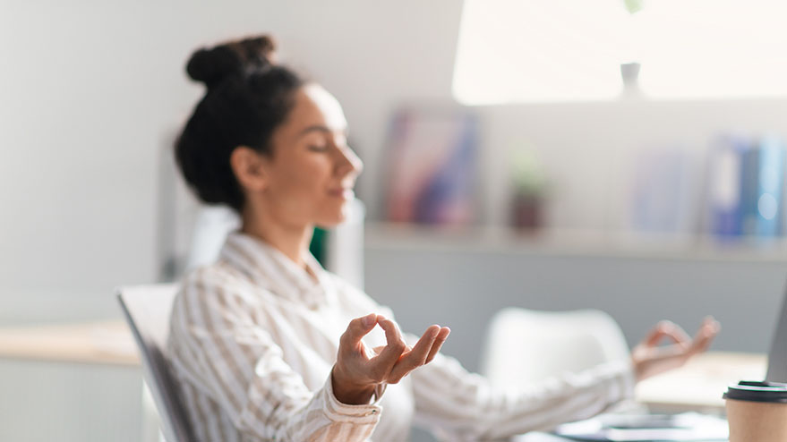 Eine Frau sitzt am Schreibtisch, sie hat die Augen geschlossen, ihre Arme und Hände in einer entspannenden, meditierenden Yoga-Haltung 