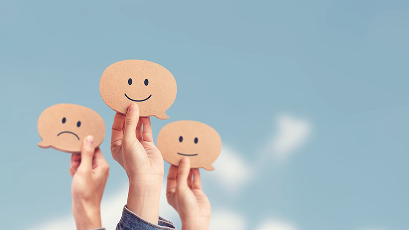 Blue sky with white clouds. Three hands hold up cardboard speech bubbles. Emojis with different facial expressions (happy, neutral, unhappy) on them.