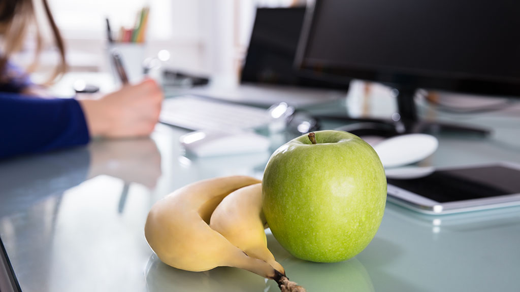A desk. On it lie two bananas and a green apple. In the background on the left is the hand of a person writing. On the right is a monitor, a smartphone lies next to the fruit.