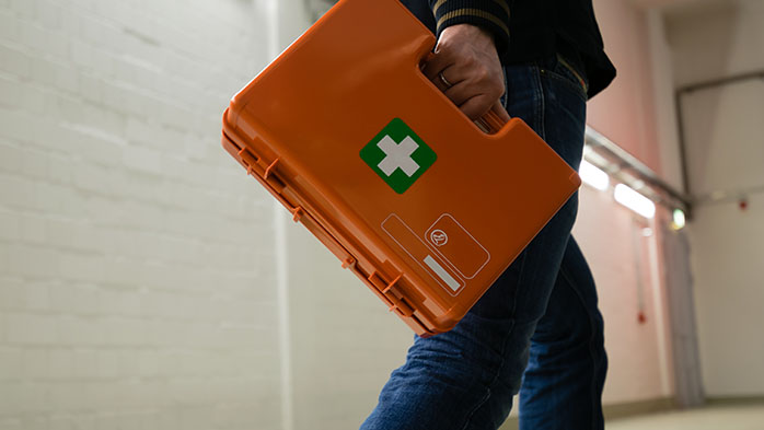 Nahaufnahme, Anschnitt: Eine Person trägt einen orangen Erste-Hilfe-Koffer. Sie trägt Jeans und läuft durch eine Werkhalle.