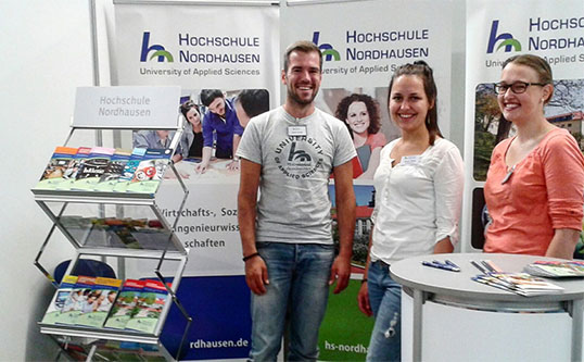 Three people stand friendly smiling at the booth of the HS Nordhausen, behind them posters and flyers