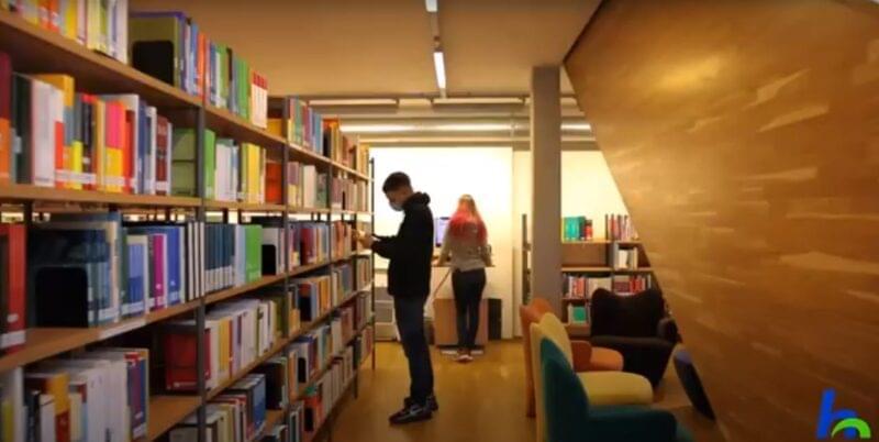 Blick ins Innere der Bibliothek der Hochschule Nordhausen. Links ein langes Bücherregal. In der Mitte steht ein junger Mann, er trägt eine Maske und hält ein geöffnetes Buch in den Händen. Eine junge Studierende steht im Hintergrund, sie arbeitet an einem PC.