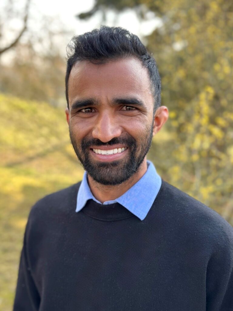 Portrait Vinod Kumar Banoth - Er trägt ein blaues Hemd unter einem dunklen Pullover, hat dunkle Haare und einen Bart. Er lächelt freundlich. Hinter ihm verschwommen Bäume und andere Pflanzen.