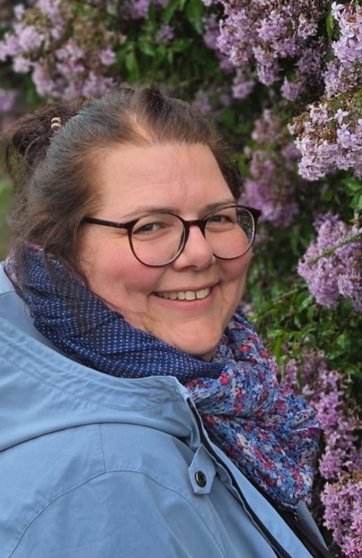 Portrait Jantje Samtleben - eine Frau mit Brille und blauer Jacke lächelt freundlich in die Kamera. Im Hintergrund sind rosa blühende Pflanzen.