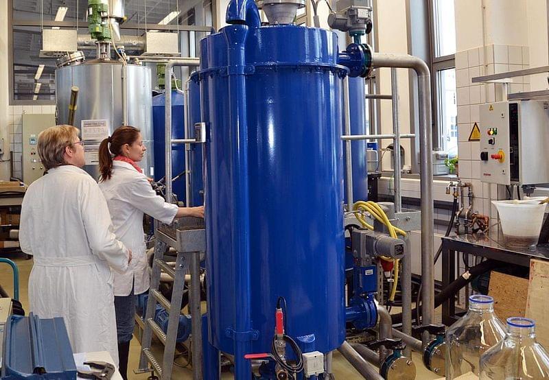 Zwei Personen in weißen Laborkitteln arbeiten an einer großen blauen Industriemaschine in einem Laborumfeld. Verschiedene technische Anlagen und Rohre sind im Hintergrund sichtbar.