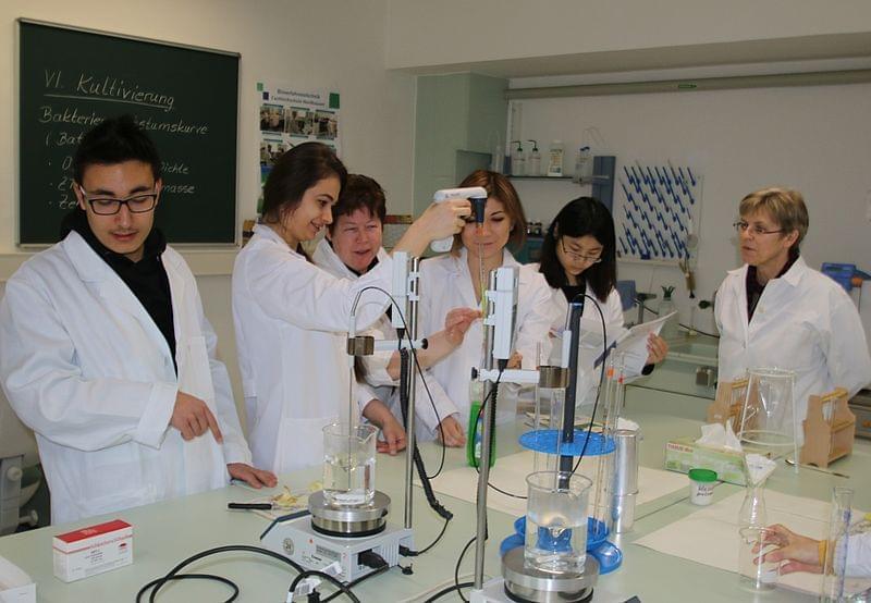 Eine Gruppe von fünf Personen in weißen Laborkitteln steht an einem Labortisch in einem Raum mit einer Tafel im Hintergrund. Sie arbeiten gemeinsam an einem Experiment, wobei eine Person in der Mitte eine Pipette hält und Flüssigkeit in ein Gerät gibt. Andere Laborutensilien wie Flaschen und Messgeräte sind auf dem Tisch verteilt.