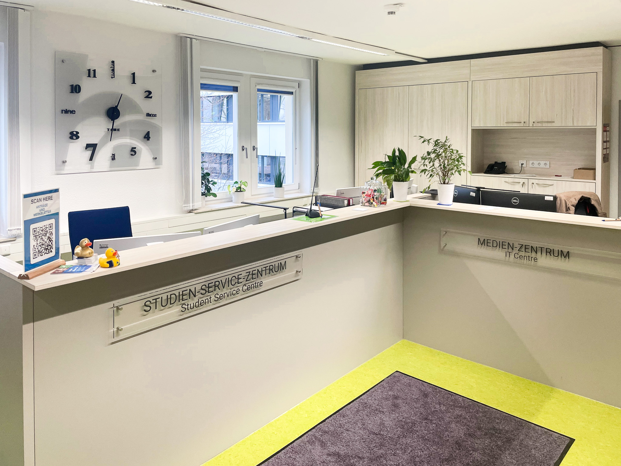 Empfangsbereich des Studien-Service-Zentrums und Medienzentrums der Hochschule Nordhausen mit hellen Möbeln, einem modernen Wandschrank, zwei Fenstern, Pflanzen und einer auffälligen Designuhr an der Wand. Auf dem Tresen stehen ein QR-Code-Schild und zwei kleine Quietscheenten.