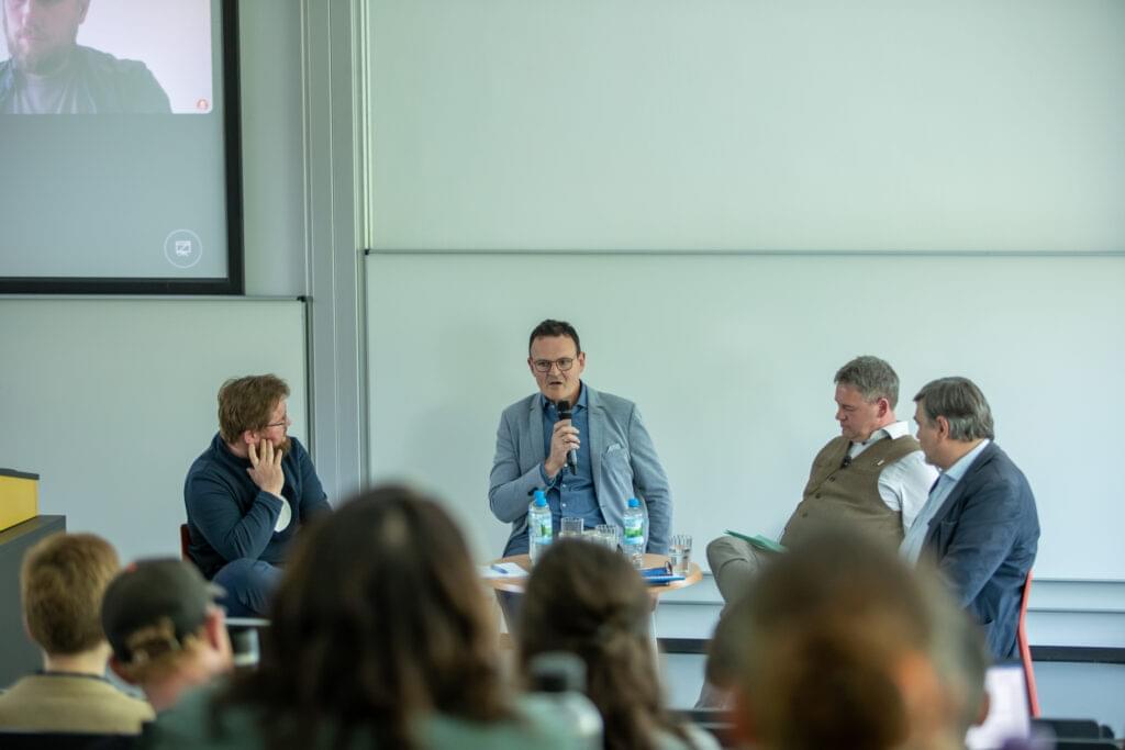 Eine Podiumsdiskussion in einem Seminarraum. Vier Männer sitzen auf Stühlen vor einem Publikum und diskutieren. Prof. Jörg Wagner hält ein Mikrofon und spricht, während die anderen aufmerksam zuhören. Im Hintergrund ist eine Leinwand mit dem Bild eines weiteren Teilnehmers, der virtuell an der Diskussion teilnimmt. Auf dem Tisch vor den Rednern stehen Wasserflaschen und Gläser. Das Publikum ist teilweise sichtbar.