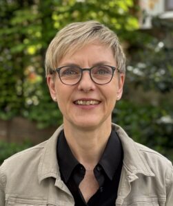 Portrait Dr. Anke Buschmann - eine Frau mit kurzen blonden Haaren. Sie trägt eine Brille und eine dunkle Bluse unter einer hellen Jacke. Sie lächelt freundlich.