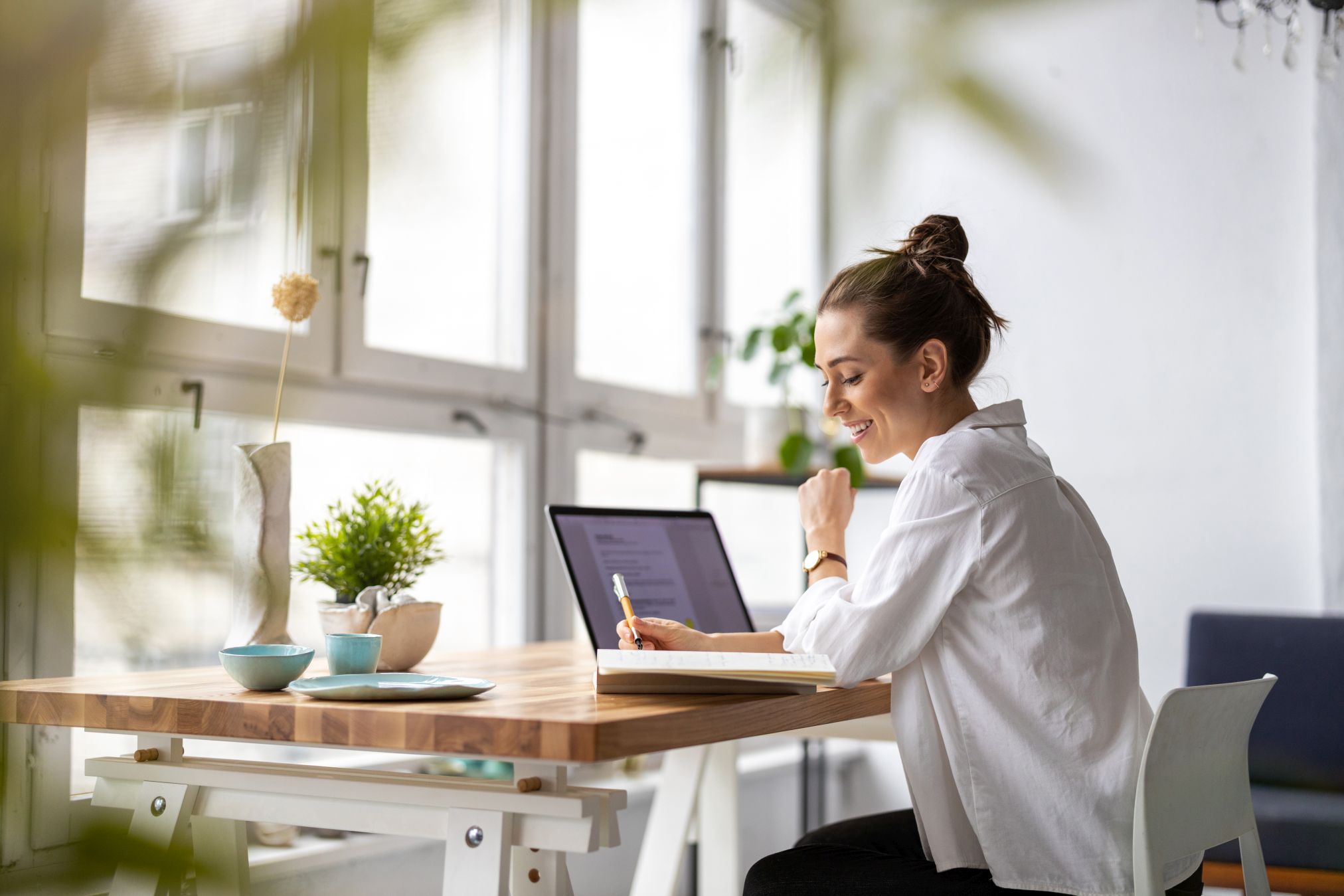 Young woman sits smiling at a bright desk with laptop and notebook, writing with concentration and working in a modern, cosy home office with plants and natural light.