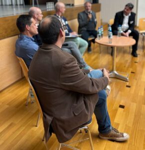 Men in business attire sit in a semi-circle and discuss on a stage during a panel discussion; bottles of water are on the table.