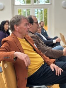 Middle-aged men sit attentively in the audience of an event in a bright hall with large windows; focus on participants in brown and orange jackets.