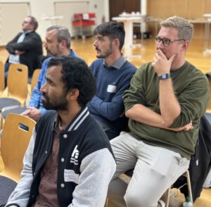 Several young men are sitting next to each other in the audience, one is wearing a Nordhausen University of Applied Sciences jacket with a "Banoth" name tag, they are looking attentively ahead.