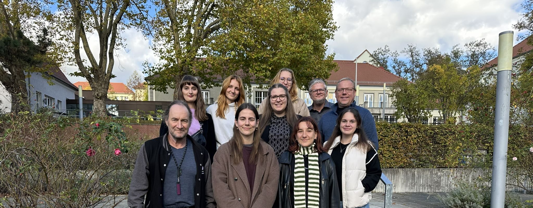 Group photo with ten people, taken outdoors on a staircase in front of autumn-coloured trees and buildings with red roofs. Five people are standing in the first row, including three women and two men; five more people are standing in the second row. They are all smiling at the camera, the atmosphere is friendly and relaxed.