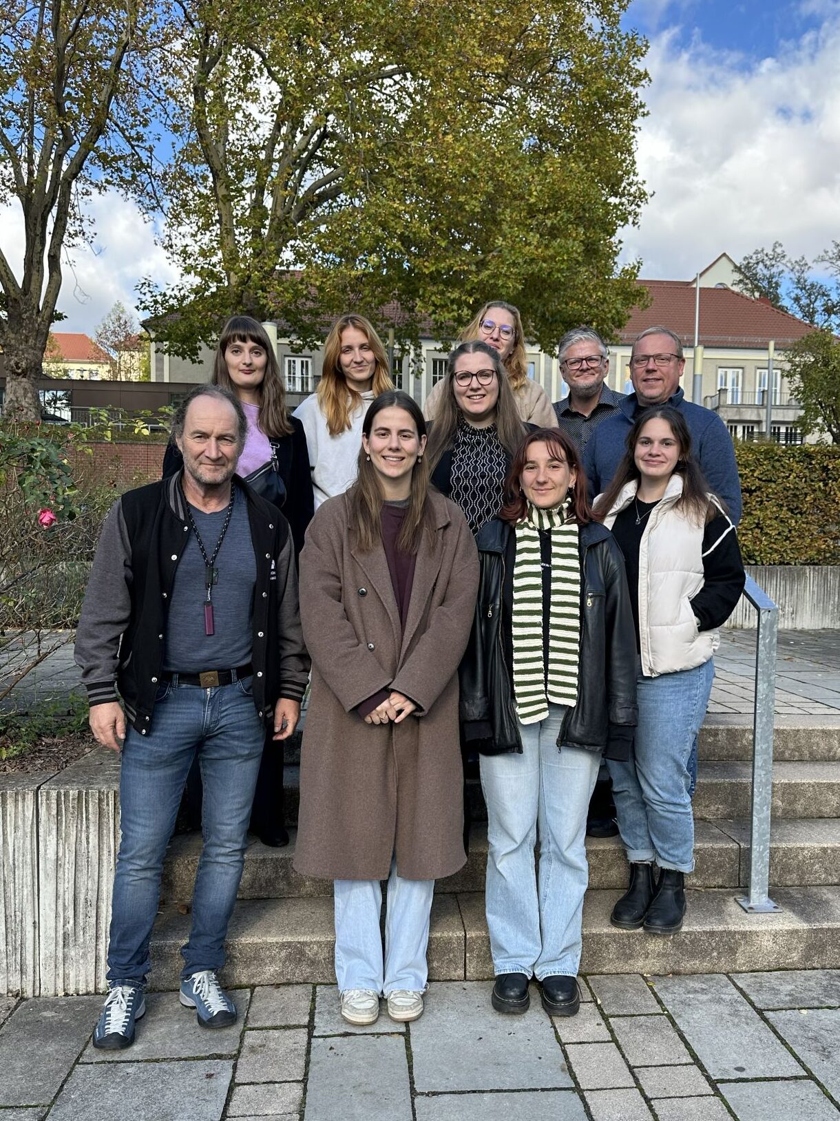 Group photo with ten people, taken outdoors on a staircase in front of autumn-coloured trees and buildings with red roofs. Five people are standing in the first row, including three women and two men; five more people are standing in the second row. They are all smiling at the camera, the atmosphere is friendly and relaxed.