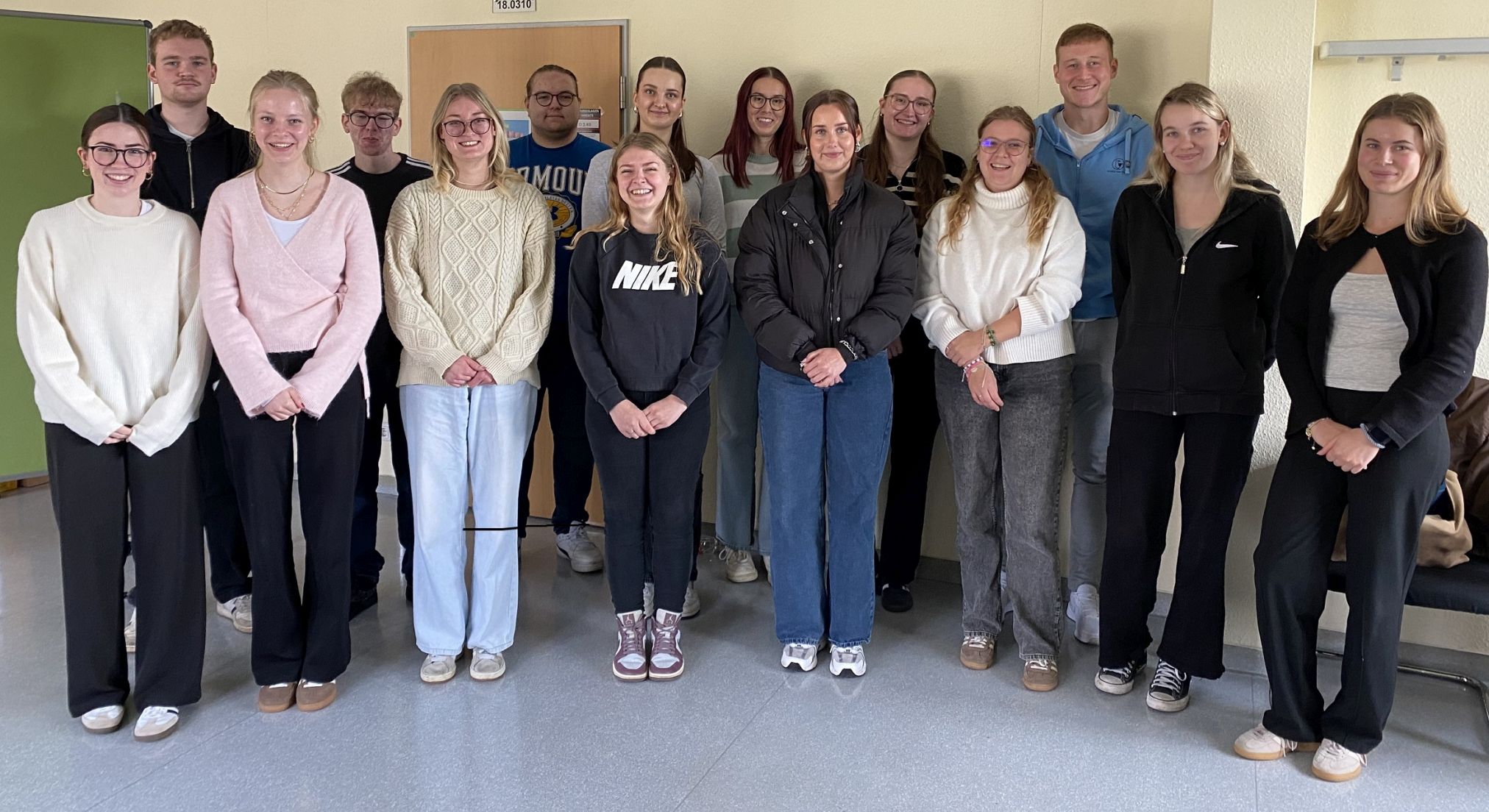 Gruppenfoto von 15 jungen Erwachsenen, die lächelnd in zwei Reihen in einem hellen Flur stehen. Sie tragen überwiegend Freizeitkleidung und blicken direkt in die Kamera – ein Willkommensmoment zum Studienstart.