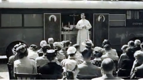 Black and white photograph of a chapel car of the aid organisation „Church in Need“ after the Second World War; a priest stands in front of the open door and speaks to a large number of people sitting in front of it.