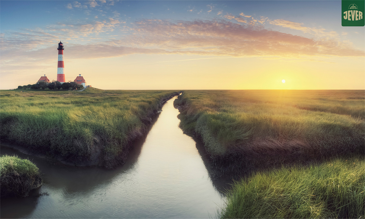 Werbeaufnahme der Biermarke Jever.
Das Foto visualisiert die Markenwerte durch eine typisch norddeutsche Szenerie:
Landschaft: Im Vordergrund zieht sich ein Priel (Wasserlauf) durch das grüne Marschland des Wattenmeers.
Architektur: Im linken Hintergrund steht der markante, rot-weiß gestreifte Leuchtturm Westerheversand mit seinen zwei Nebengebäuden.
Lichtstimmung: Die Sonne steht tief am Horizont und taucht den Himmel in warme Gelb- und Orangetöne.
Branding: In der oberen rechten Ecke ist dezent das grüne Logo der Biermarke JEVER eingeblendet.