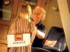Das Bild fängt einen herzerwärmenden Moment in warmen Goldtönen ein. Ein junges Mädchen mit Zöpfen und einem karierten Kleid steht mit dem Rücken zum Betrachter und hält eine Merci-Packung hinter ihrem Rücken oder überreicht sie gerade. Ihr Gegenüber ist eine ältere Frau mit blondem Haar, die das Kind liebevoll und freudig anlächelt. Die Atmosphäre wirkt häuslich und vertraut.