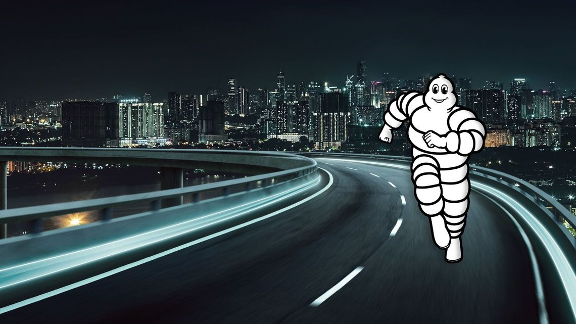 Die Grafik zeigt eine moderne Autobahnbrücke bei Nacht, deren Fahrbahnmarkierungen durch Langzeitbelichtung als helle Lichtstreifen erscheinen. Im rechten Vordergrund rennt die ikonische Werbefigur „Bibendum“ (das Michelin-Männchen) mit einem freundlichen Lächeln in Richtung des Betrachters. Im Hintergrund ist die hell erleuchtete Skyline einer Großstadt bei Nacht zu sehen.