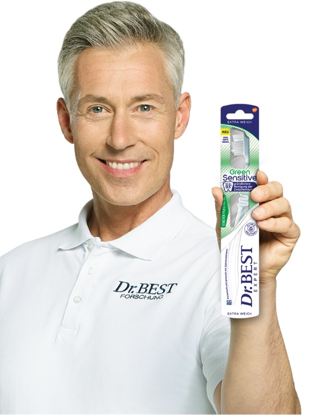 Das Bild zeigt einen grauhaarigen Mann im Porträt, der ein weißes Polohemd mit der Aufschrift „Dr.BEST FORSCHUNG“ trägt. Er lächelt in die Kamera und hält in seiner rechten Hand eine originalverpackte Zahnbürste hoch. Die Verpackung ist als „Green Sensitive“ und „EXTRA WEICH“ gekennzeichnet. Ein Siegel auf der Packung weist das Produkt als „KLIMA NEUTRAL“ aus.