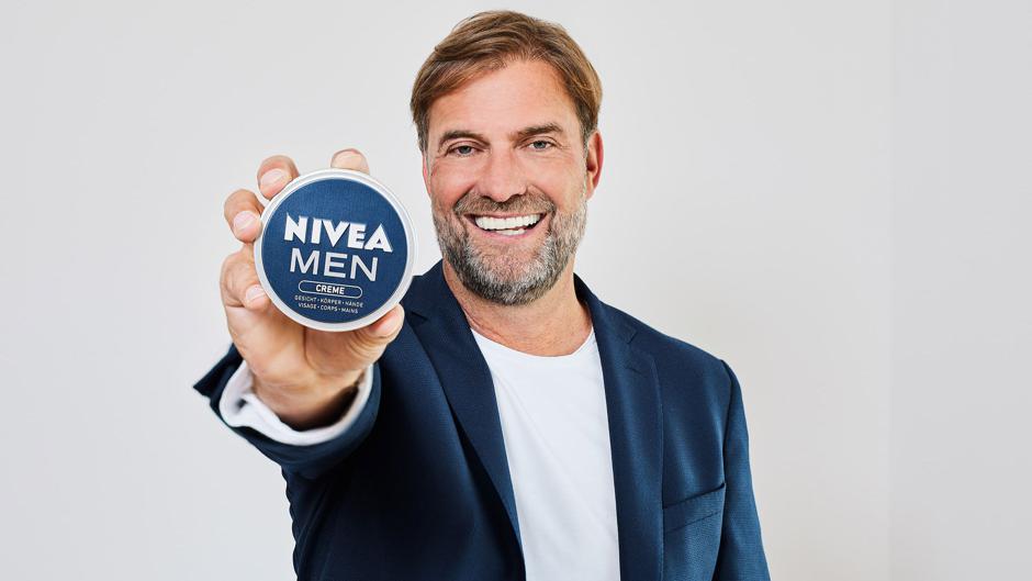 Das Bild zeigt Jürgen Klopp in einem dunkelblauen Sakko über einem weißen T-Shirt vor einem hellgrauen Hintergrund. Er blickt lachend in die Kamera und hält in seiner ausgestreckten rechten Hand eine blaue Dose „NIVEA MEN CREME“. Die Dose ist so positioniert, dass der Produktname für den Betrachter zentral und deutlich lesbar ist.
