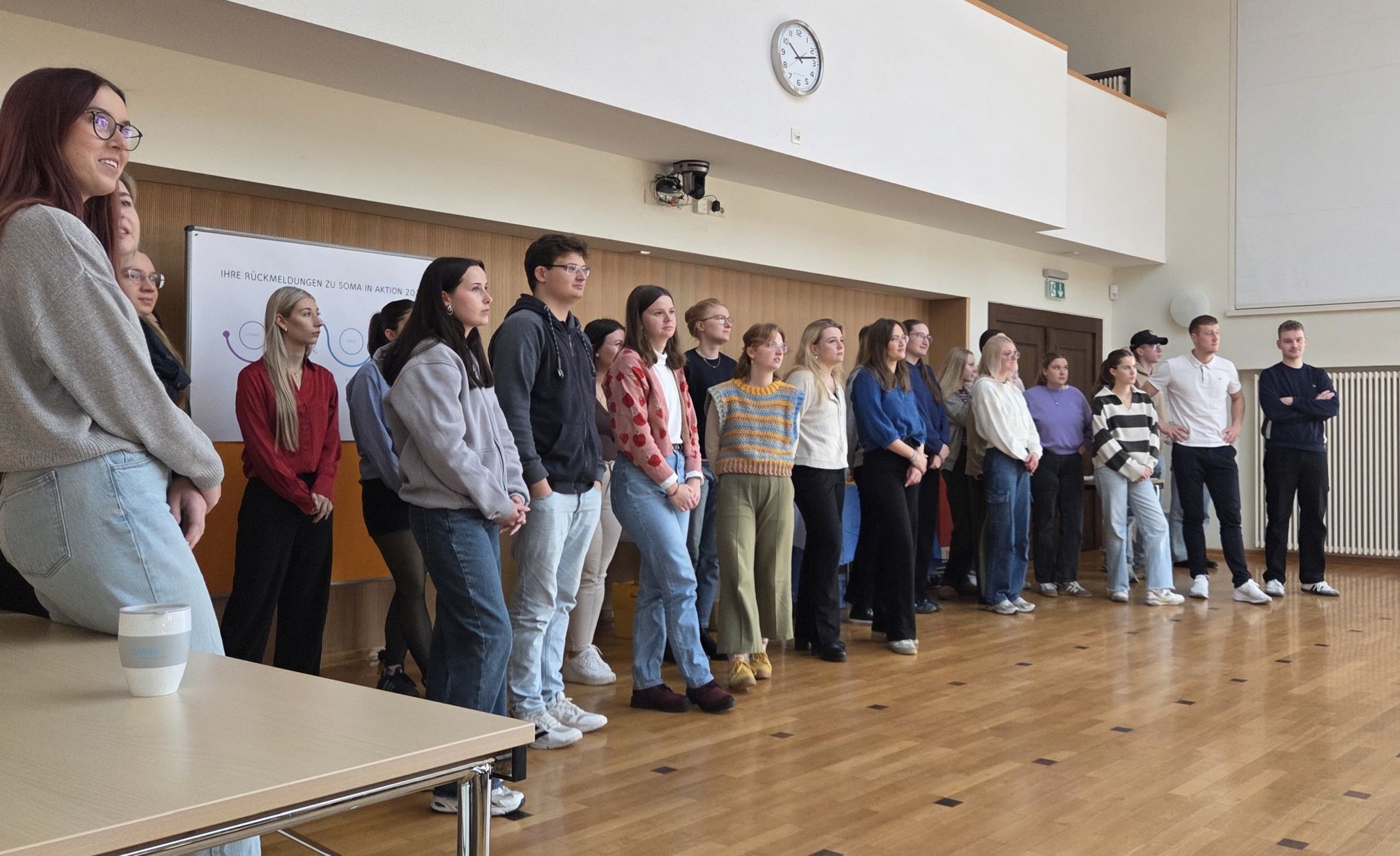 Eine große Gruppe junger Erwachsener steht nebeneinander im Audimax vor einer weißen Wand mit Projektor. Im Hintergrund ist ein Schild mit dem Schriftzug „SOMA in Aktion“ zu sehen.