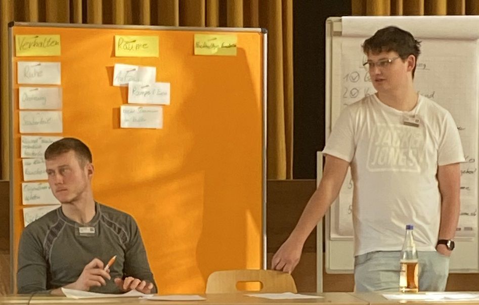 Two young men stand and sit in front of a pinboard with notes organised into categories such as „Behaviour“, „Routine“ and „Sustainable approach“. One of them is wearing a Jack & Jones T-shirt, the other is sitting with a pen in his hand.