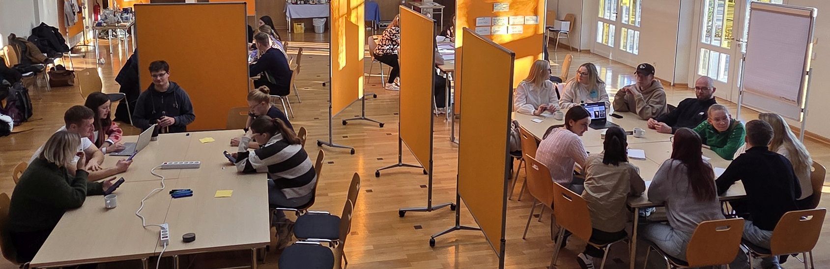 Several groups of young people are working at long tables in a bright room with mobile pinboards. Laptops, pens and notepads are spread across the tables.
