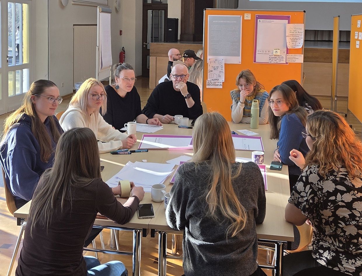 Elf Personen sitzen im Kreis um einen Tisch und diskutieren, einige mit Stiften und Heften. Im Hintergrund hängen orange Pinnwände mit Aufgabenstellungen und Ergebnissen.