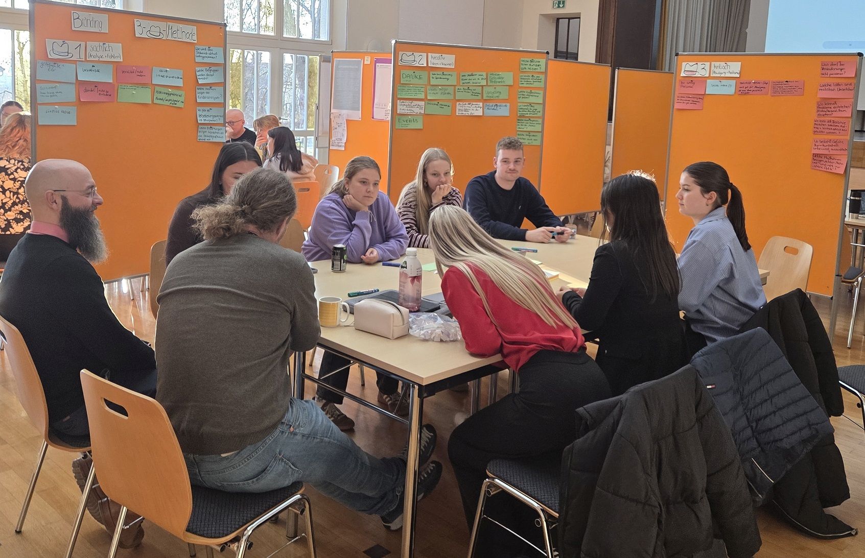 A group of students sits at a table in front of coloured moderation cards with headings such as „Bundling“ and „3-Cs method“. The atmosphere is focused and collaborative.