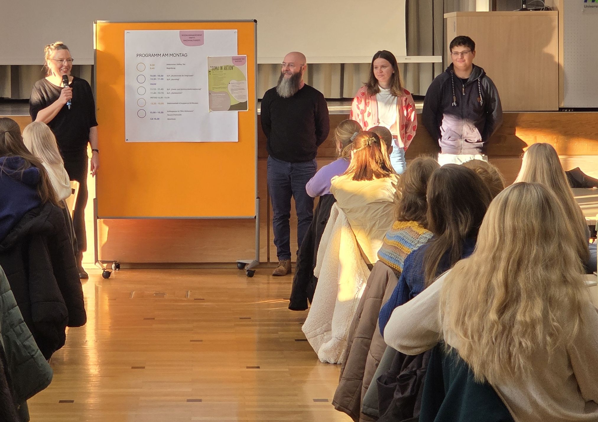 Four people stand in front of a group of seated participants. A programme poster with the day's schedule is shown on the left. A woman speaks into a microphone.
