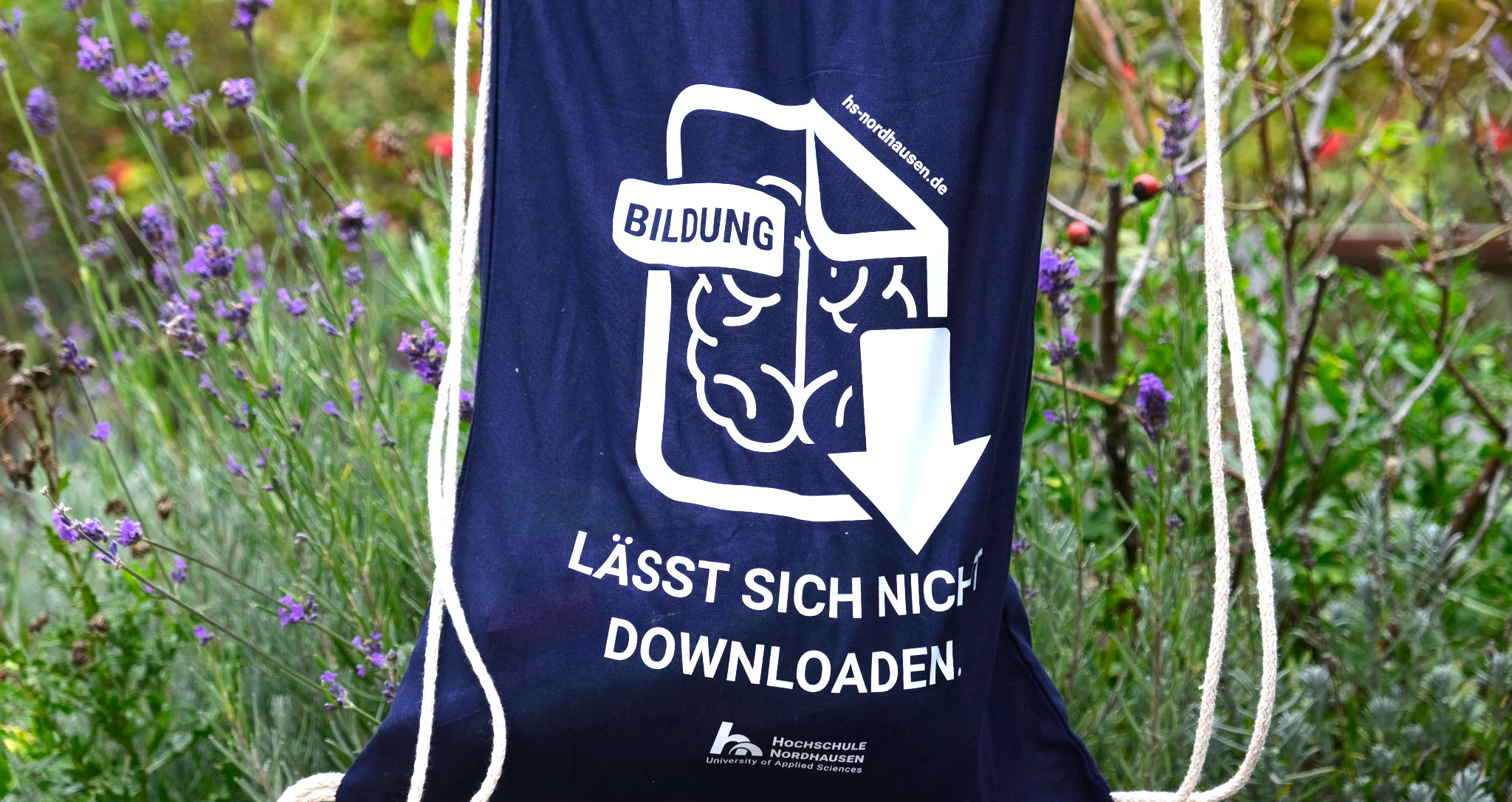 Ein blauer Turnbeutel mit der Aufschrift "Bildung lässt sich nicht downloaden". Darunter das Logo der Hochschule Nordhausen. Im Hintergrund der grüne Campus.